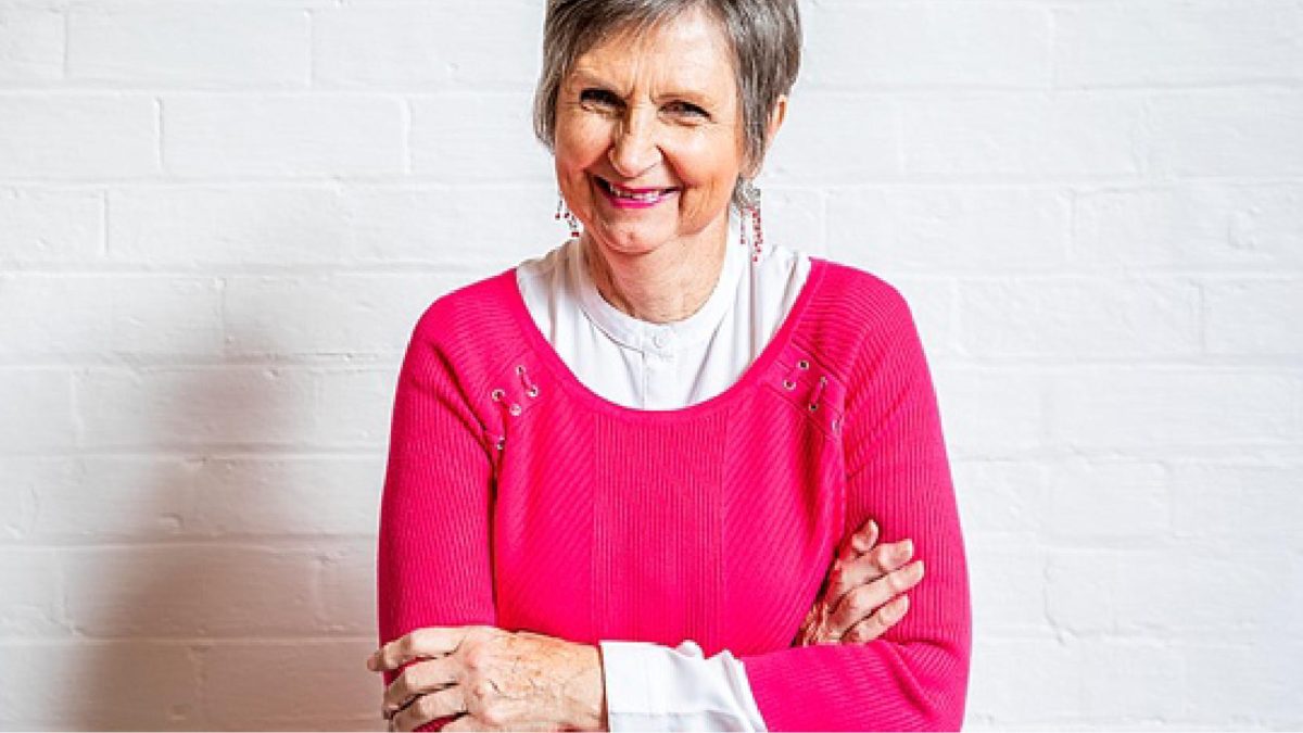 Older lady in a pink sweater with her arms crossed smiling.