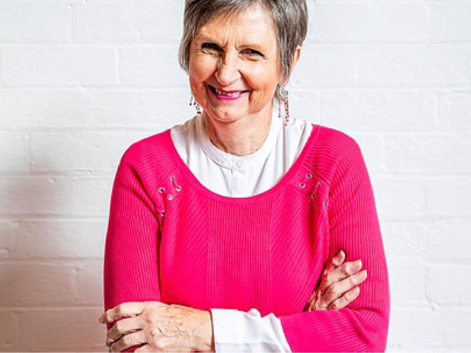 Older lady in a pink sweater with her arms crossed smiling.